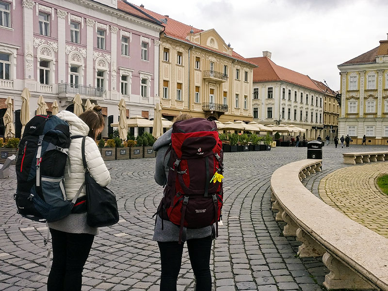 Two girls with large backpacks on a square that is lined with old, colourful buildings.