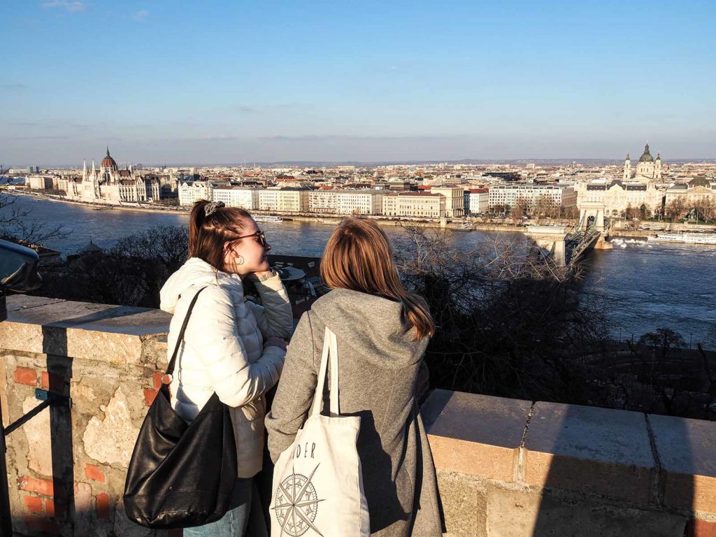 Two girls in the front looking to the horizon. In the middle a river and int he background the city of Budapest.