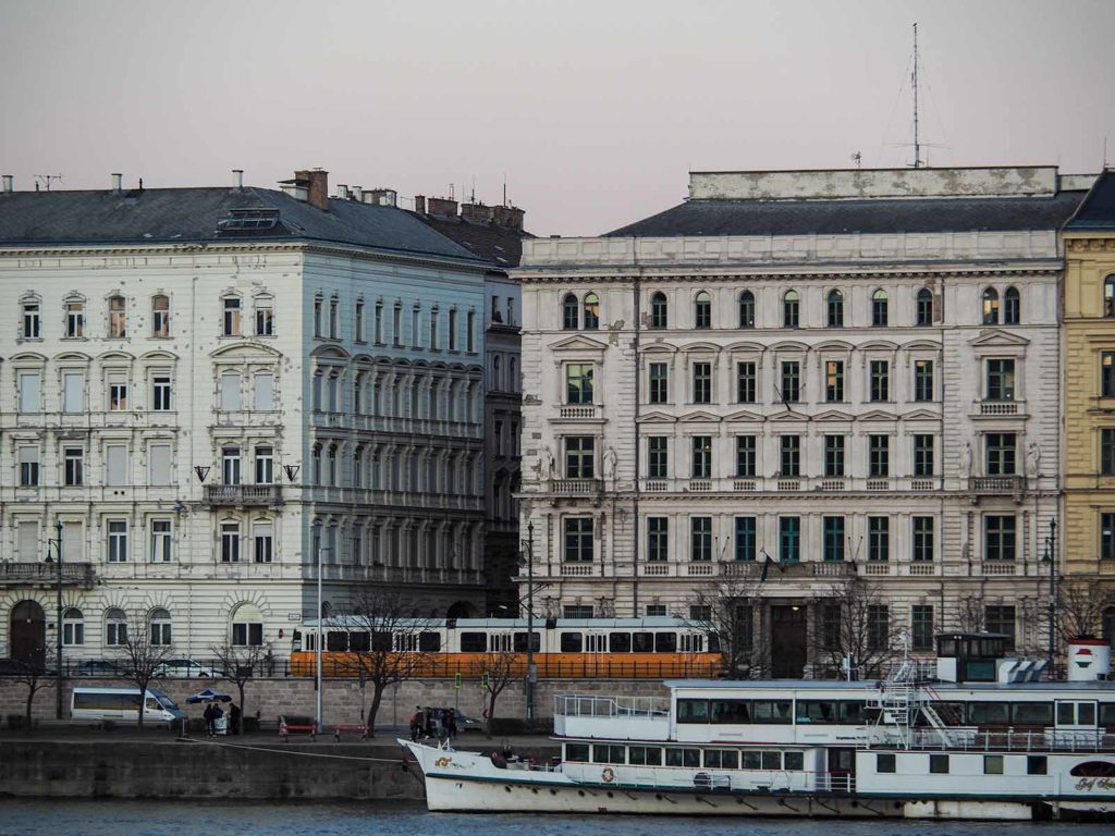 In the front, a small stretch of the river Danube, behind the river a orange tram passing by old houses.