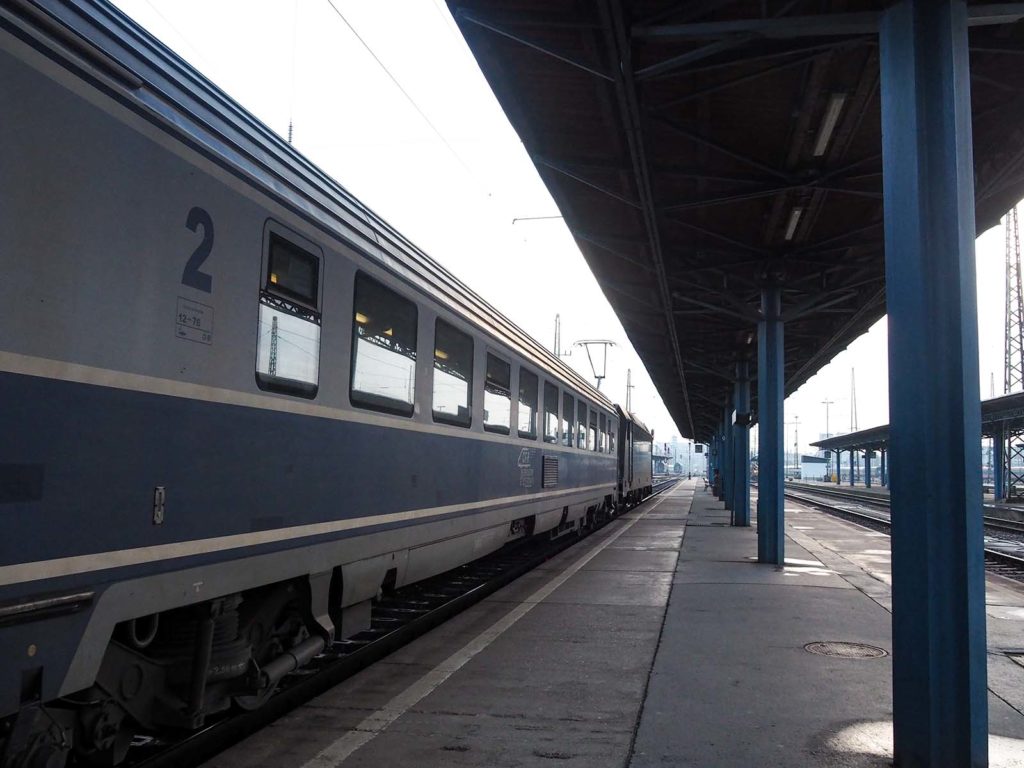 Train station in Budapest. On the left side a blue painted train that stretches to the background. In the middle and on the right side the platform.