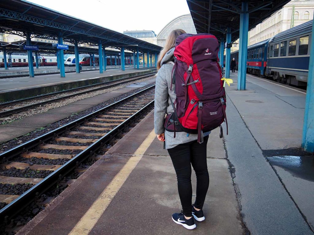 Girl with a big red backpack at the train station in Budapest. On the left side the train tracks, in the upper right corner a train.
