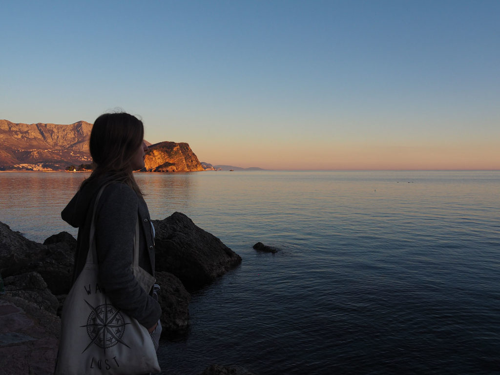 In the background the sea and some rocks. The sky is painted blue, orange and red. In the front on the left side a girl watching the sea.