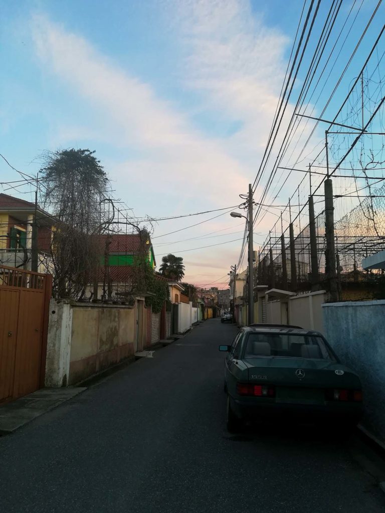 A street, on the street a car and on the sides walls and buildings behind the walls. The sky is painted orange from the sunset.
