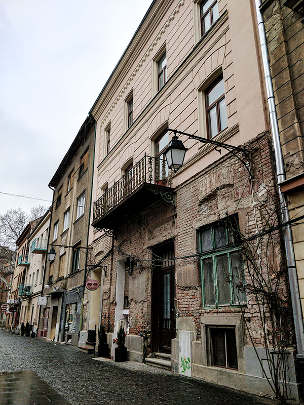 House walls of old buildings. The building in the front has a small balcony on the first floor and a brick wall on the ground floor.