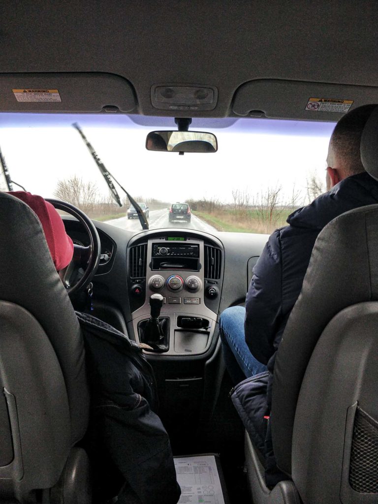 View from the back seat of a car. In the front two man.