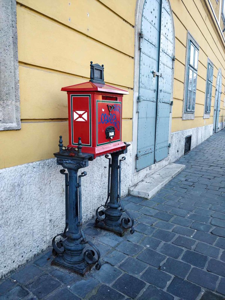 Red post box in front of a old, yellow building.